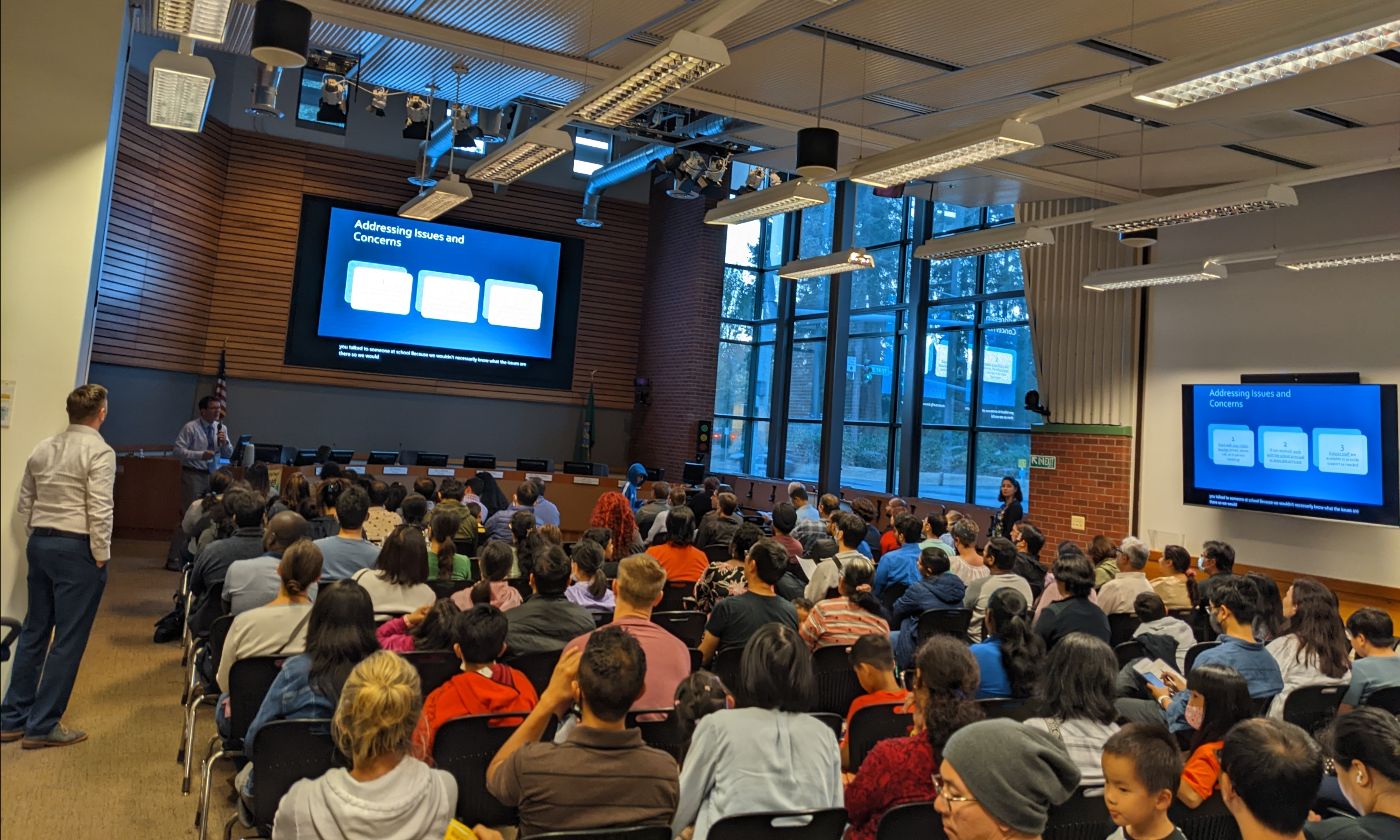 Photo of an audience of people seated in a large room, facing the front where a presentation is displayed on a large screen.