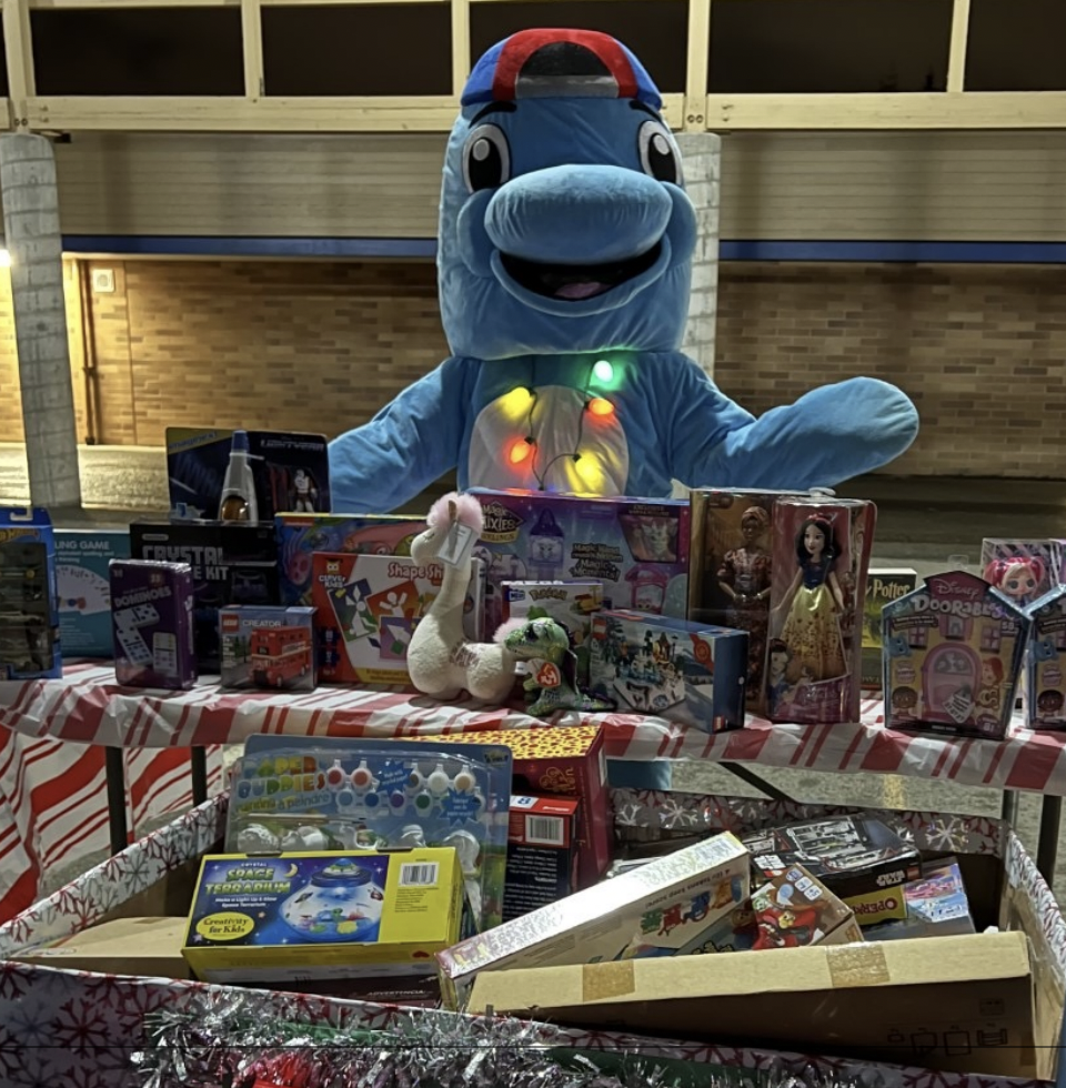 Photograph of Samantha Smith Elementary's dolphin mascot behind a Toys for Tots collection box and table filled with donated toys for kids in need at Smith PTSA's 2022 Winter Wonderland event
