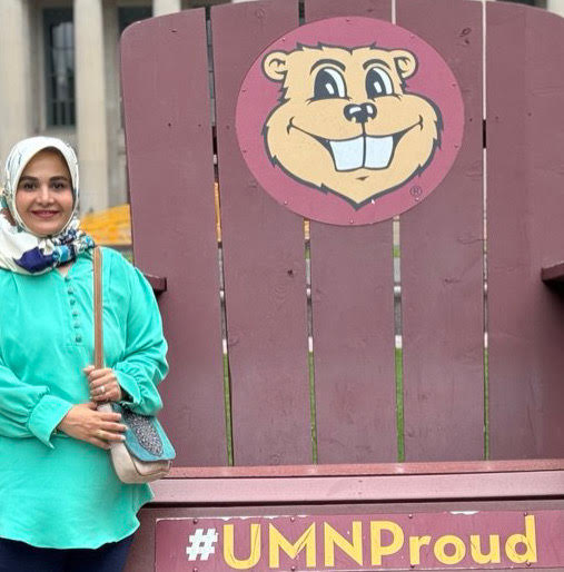A woman wearing an aqua tunic and a floral hijab stands next to an oversized adirondack chair painted with the University of Minnesota gopher mascot